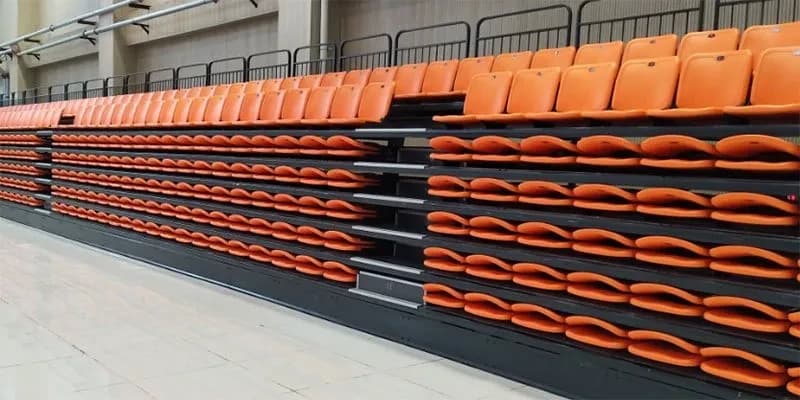 Seating brochure cover — a bank of retractable telescopic seating part-way through retraction, its orange moulded seats folded flat in stacked rows inside an indoor arena